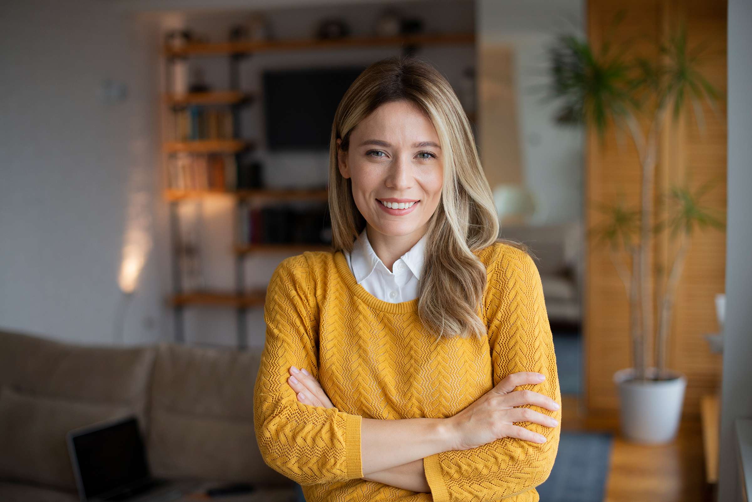 woman wearing a yellow sweater standing in her home looking straight into the camera lens with a smile on her face and her arms crossed