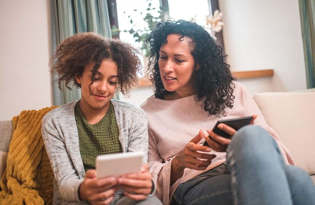 Mom and daughter sitting on couch together both holding electronic devices. Mom is looking over daughter's shoulder and smiling. Daughter is holding out device so her mom can see what she is doing.