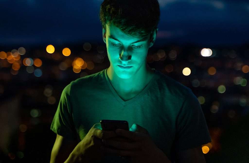 teenage boy looking at his phone in the dark with city lights behind him