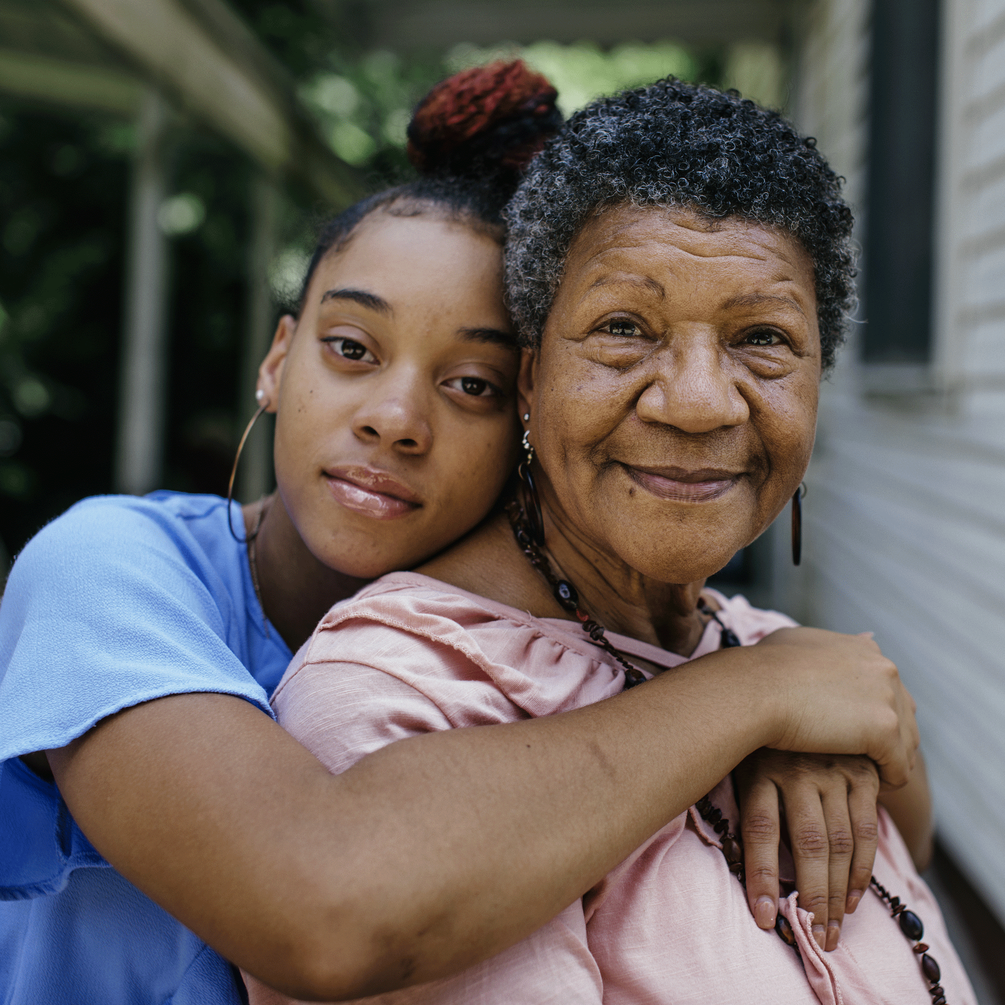 A young woman embraces an older woman from behind as they both look at the camera. The older woman smiles warmly while the younger woman has a calm, gentle expression. They are outdoors next to a house.