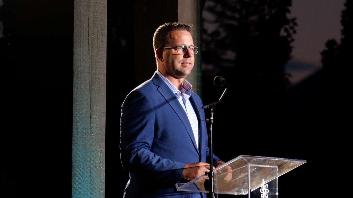 Middle aged man with brown hair, glasses, and blue suit coat with patterned button-up top standing at a podium addressing a crowd.