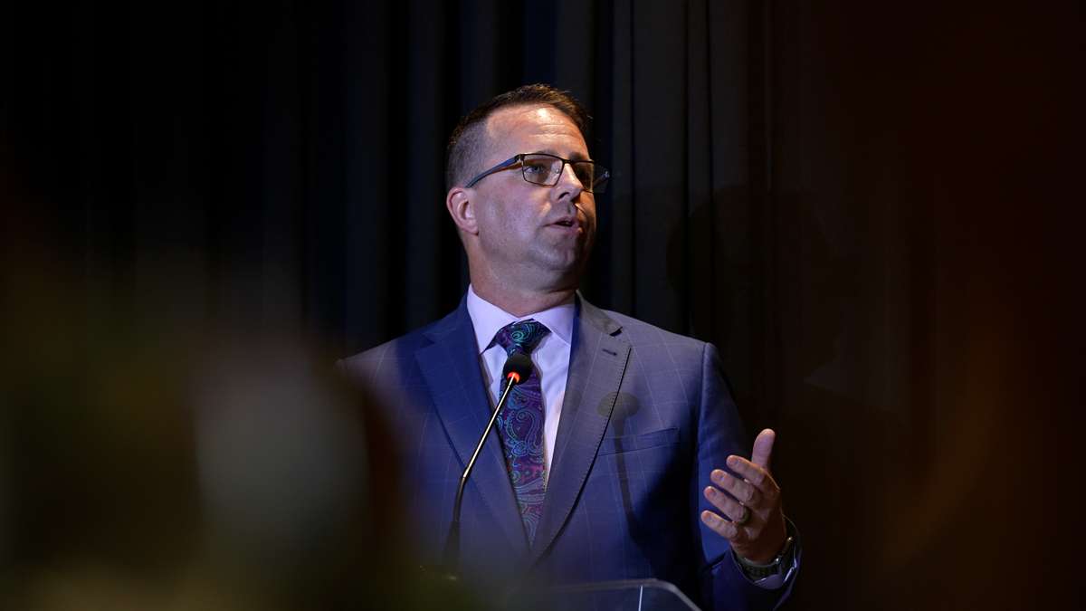 Middle aged man standing at podium speaking to an audience. He has a blue suit jacket with tie and button-up shirt, and is wearing glasses.