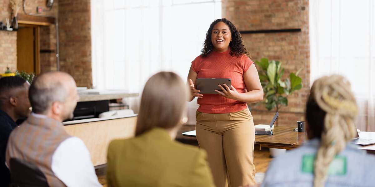 Woman standing in front of a group of people presenting. She is holding an iPad and is wearing an orange top and brown pants.