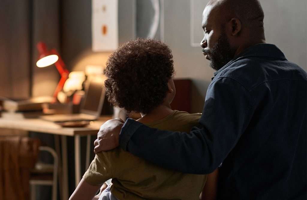 A parent sits with a child, offering comfort and support in a softly lit room. The parent has an arm around the child’s shoulders, and they appear to be having a serious, caring conversation.