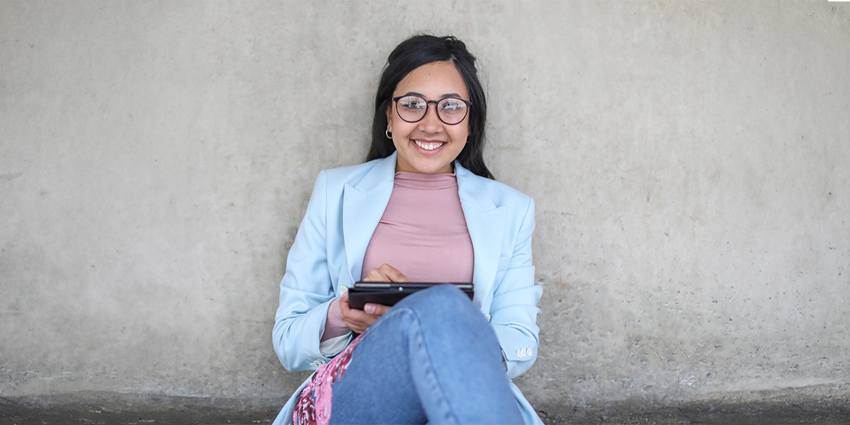 A smiling young woman wearing glasses, a light blue blazer, and jeans sits against a concrete wall while holding a tablet in her lap.