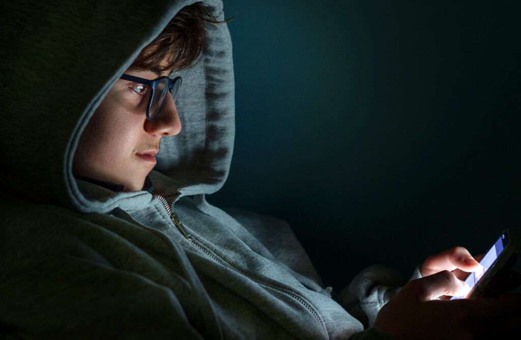 Boy wearing hoody and glasses looking a cell phone in the dark.