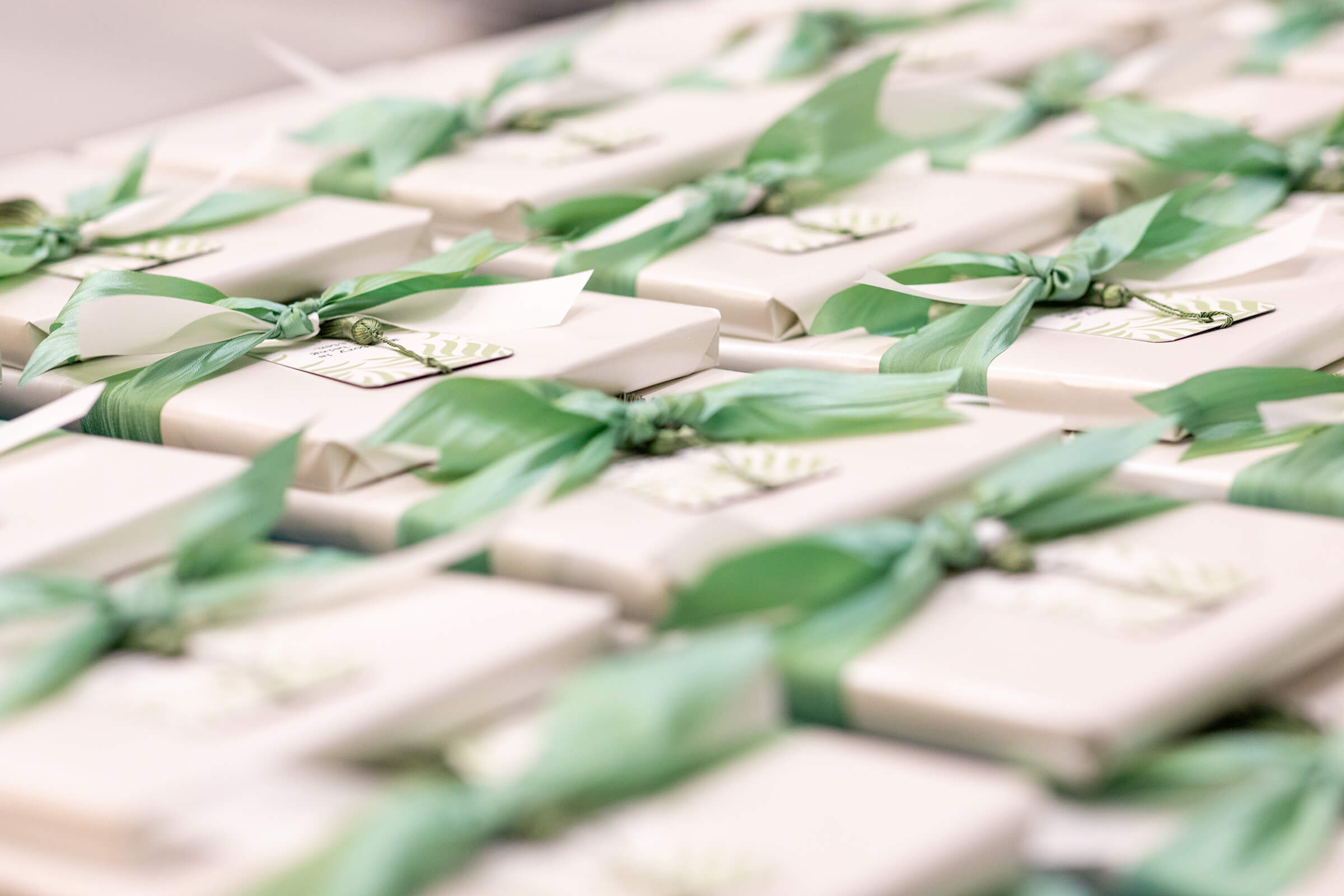 Gifts wrapped in white paper and tied with green ribbon laid across a table stacked in rows.