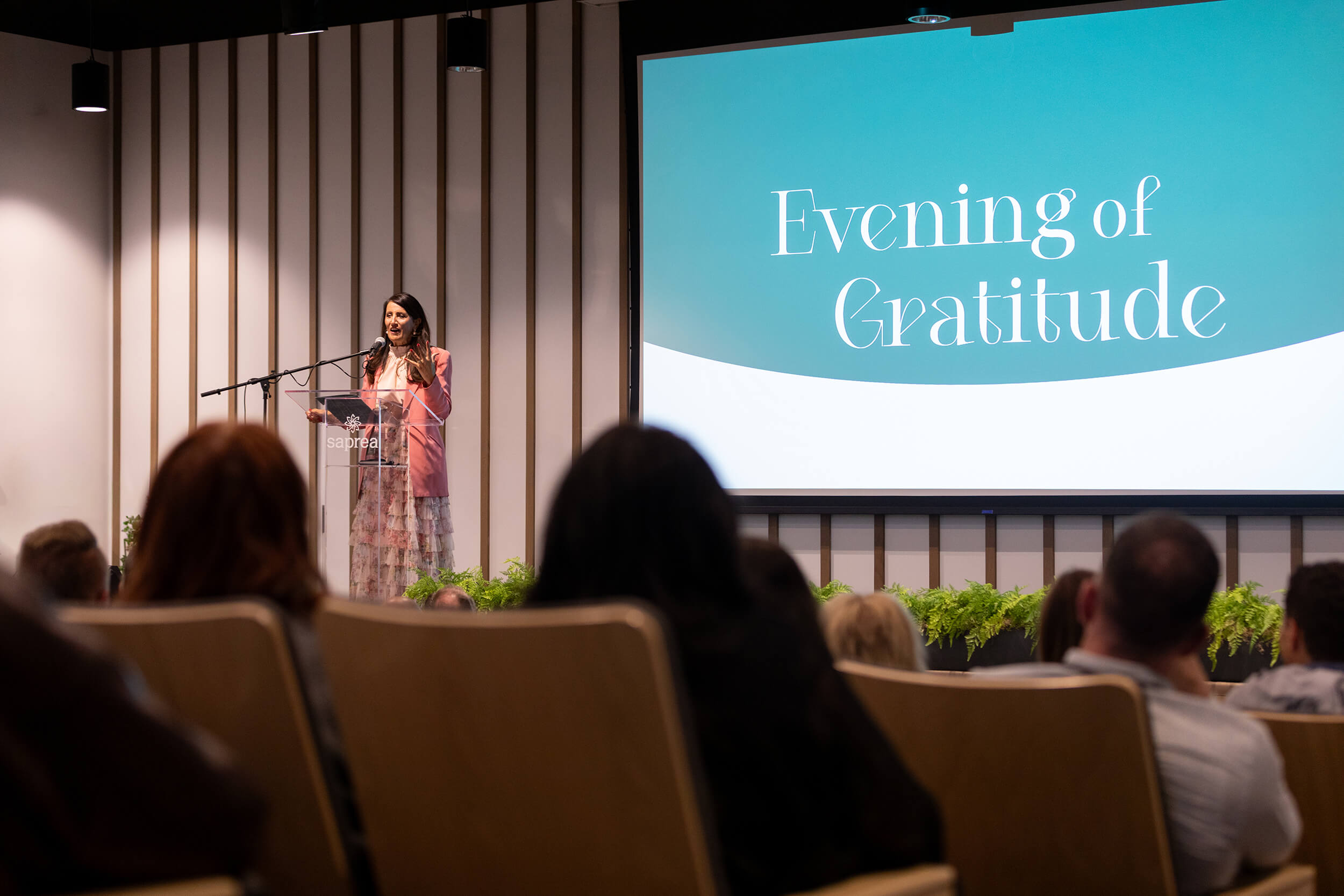 Cynthia Gambill stands at the head of the auditorium addressing the crowd for the Evening of Gratitude. She is wearing a pink dress and is standing behind a clear podium.