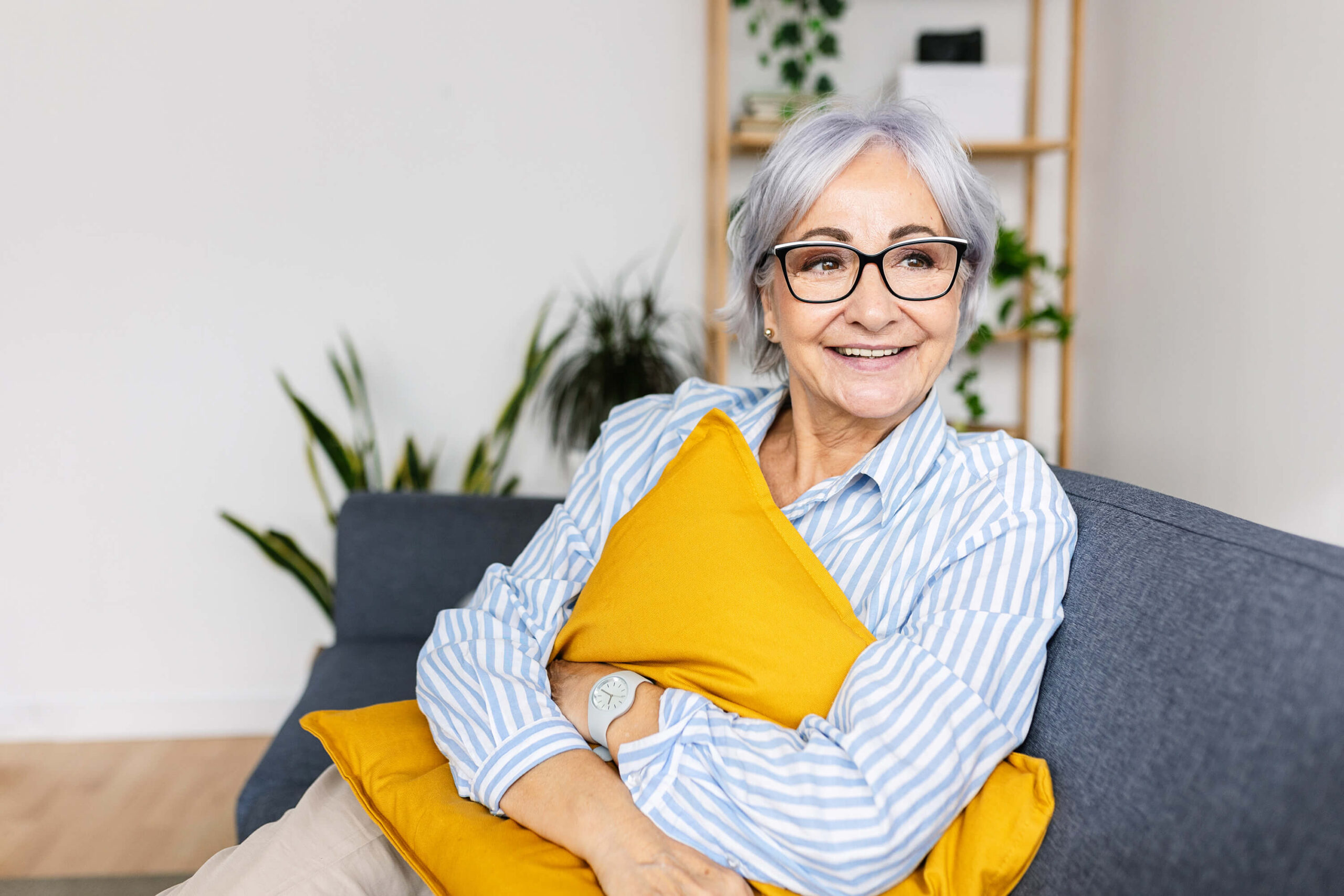 Smiling senior woman sitting on chair holding a yellow pillow up to her chest with plants in the background.