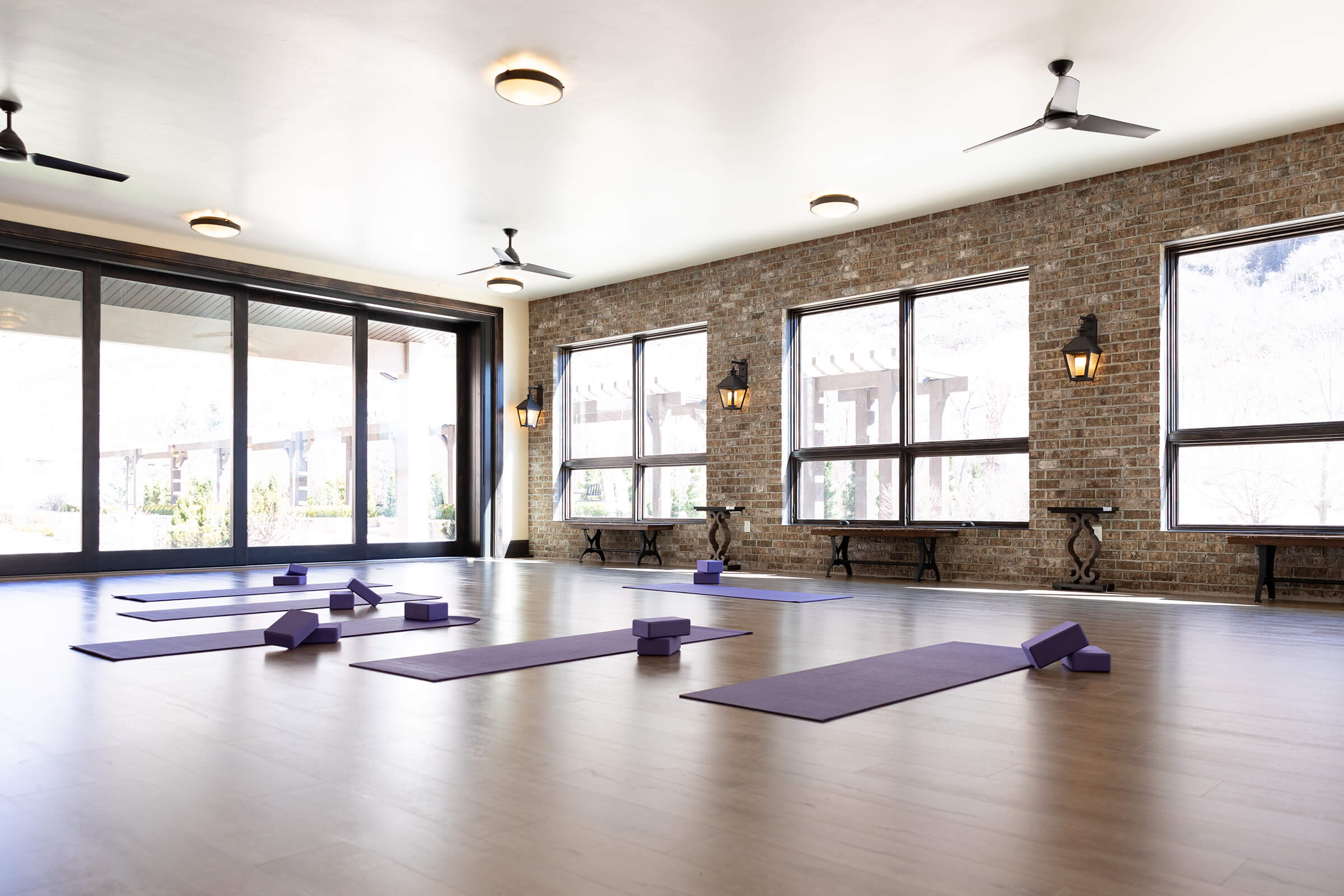 Studio space flooded in natural light with exposed brick wall and rows of open windows. There are purple yoga mats and blocks laid out across the floor.