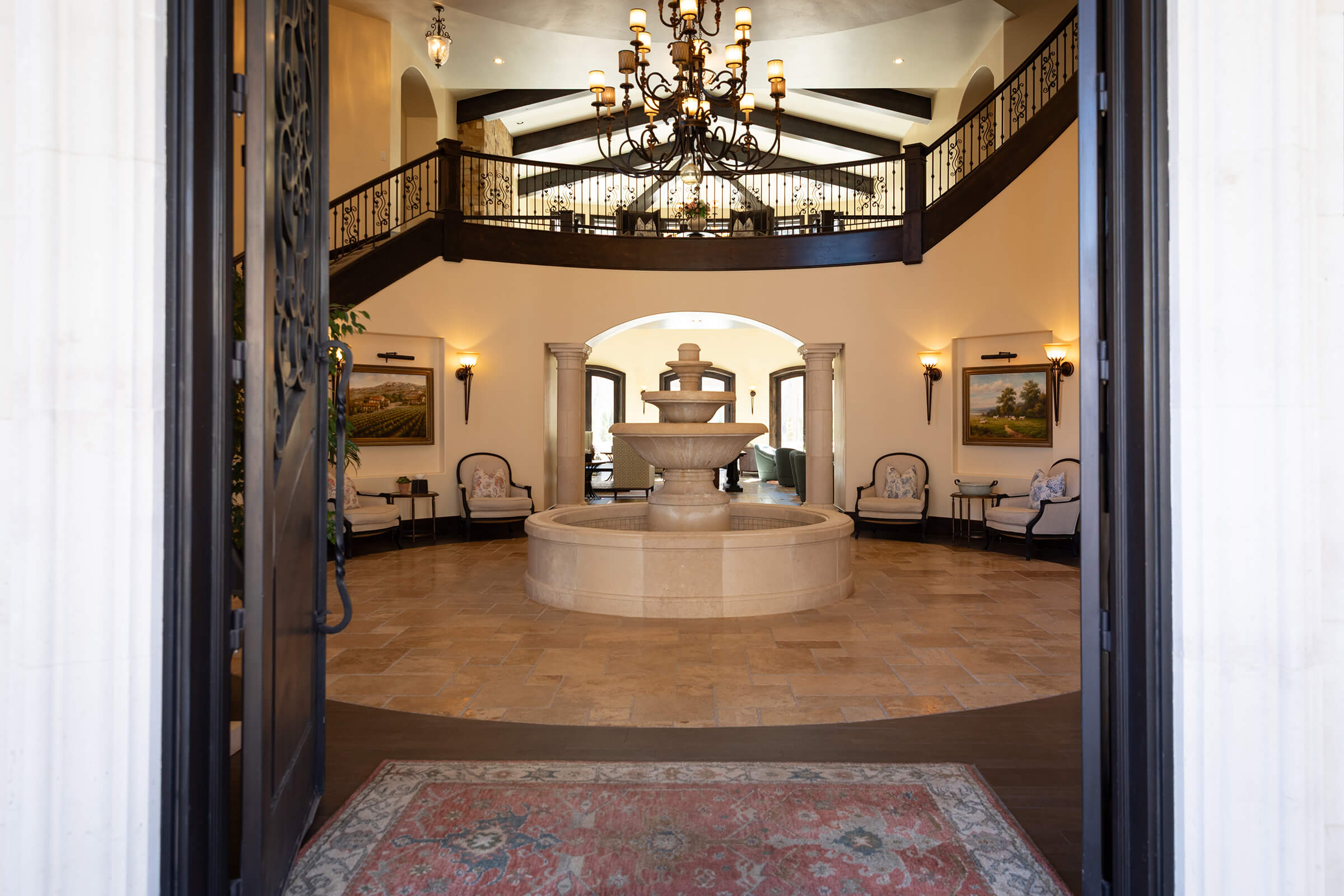 Grand entryway with limestone fountain in the middle. Room is dressed in warm lights, with swooping staircase to the second floor off to the left.