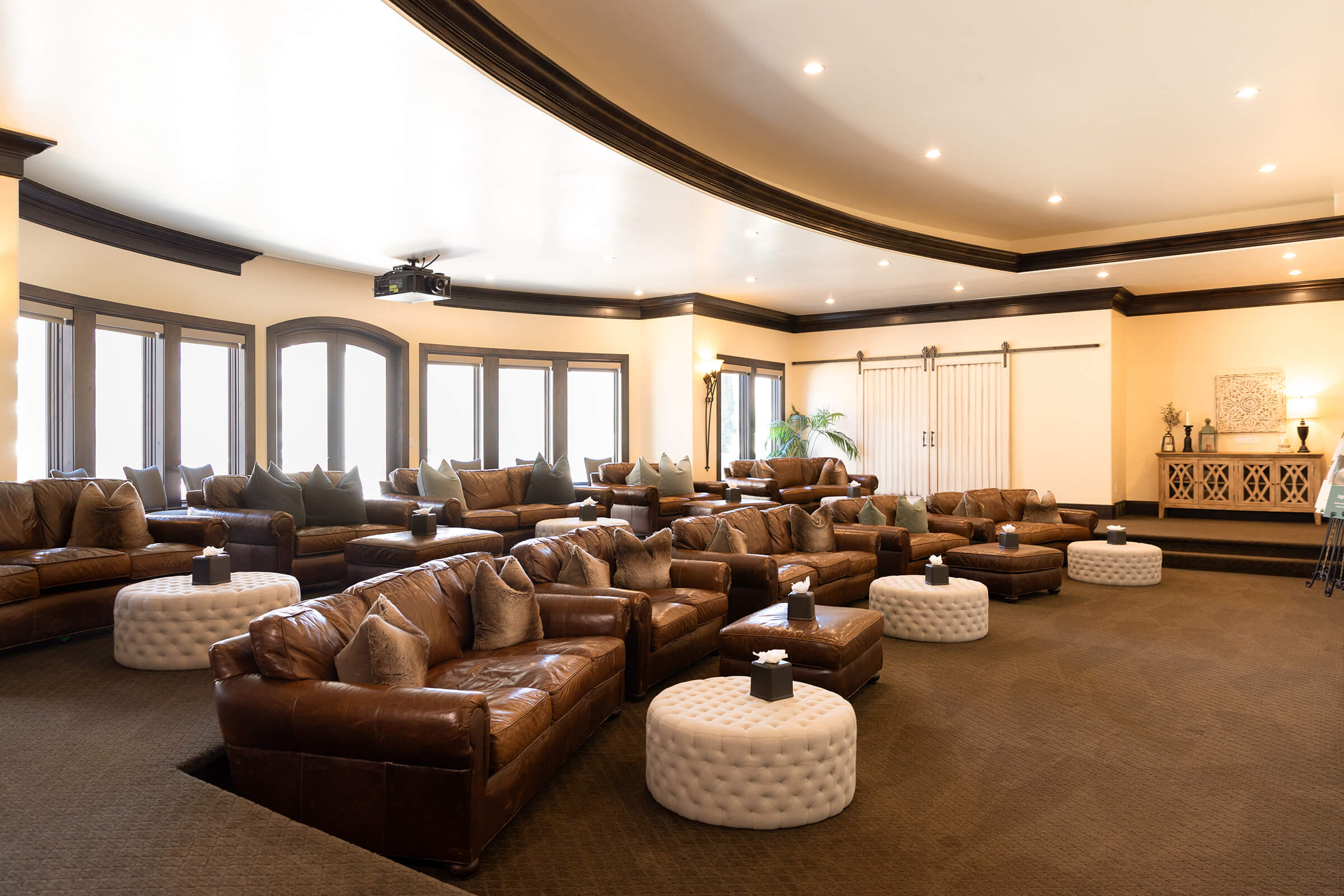 Large room with several oversized brown leather couches, fabric ottomans, and open windows that allow floods of natural light.